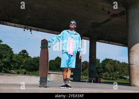 Lächelnder, modischer Mann in Casuals, der mit Longboard in der Nähe der Überführung an sonnigen Tagen steht Stockfoto