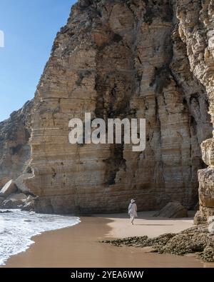 Blick von hinten auf eine Frau, die entlang des Strandes neben dramatischen Klippen an der Westspitze der Algarve geht; Sagres, Faro, Portugal Stockfoto