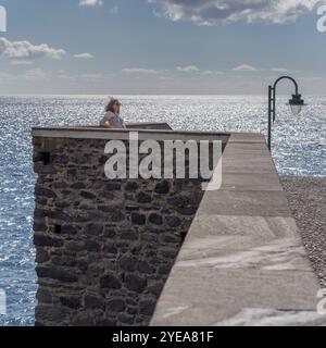 Frau steht an einer Steinmauer entlang der Uferpromenade von Ponta do Sol an der Küste Portugals, während das Meereswasser im Sonnenlicht schimmert Stockfoto