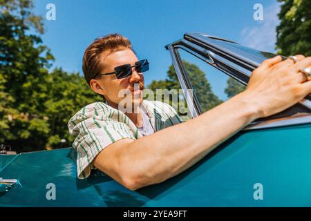 Tiefwinkelansicht eines jungen Mannes mit tätowiertem Arm, der Sonnenbrille trägt, während er im Oldtimer sitzt Stockfoto