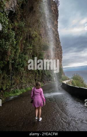 Frau steht und beobachtet einen Wasserfall, der auf einer Küstenstraße platzt, eine extrem natürliche Wasserfall-Autowaschanlage in den Felsen von Madeira, Portugal Stockfoto