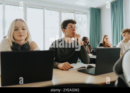 Männlicher Schüler, der bei einem blonden Mädchen sitzt und kabellose Kopfhörer am Schreibtisch im Klassenzimmer trägt Stockfoto