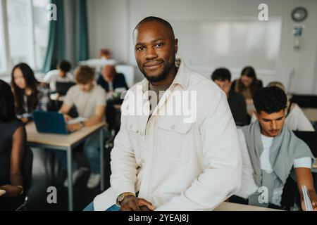 Porträt eines lächelnden männlichen Lehrers, der im Klassenzimmer von einem Schüler auf dem Schreibtisch sitzt Stockfoto