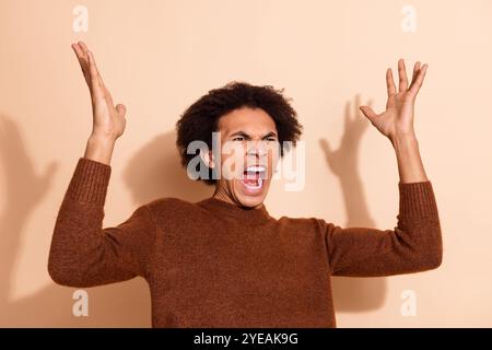 Ausdrucksstarker junger Mann im lässigen Outfit, der vor beigefarbenem Hintergrund aufgeregt die Hände hebt Stockfoto