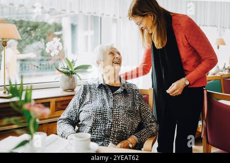 Lächelnde Frau mit Hand auf der Schulter einer glücklichen älteren Frau, die im Altersheim auf einem Stuhl sitzt Stockfoto