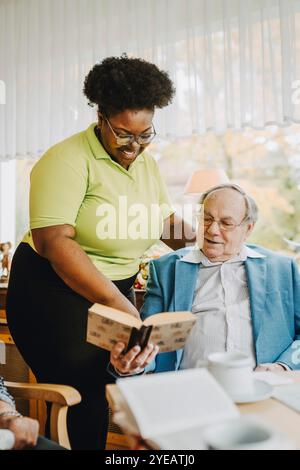 Lächelnde Pflegerin, die beim pensionierten Mann steht und im Pflegeheim Buch liest Stockfoto