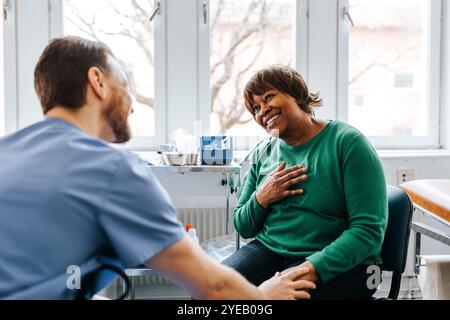 Glückliche ältere Frau mit Hand auf Brust, die mit männlichem Arzt während des Sitzens im medizinischen Untersuchungsraum spricht Stockfoto