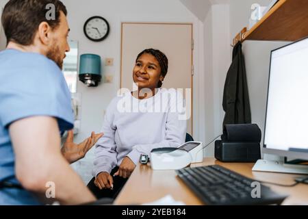 Männlicher Arzt, der junge Frauen erklärt, während er in der Nähe des Tisches im medizinischen Untersuchungsraum sitzt Stockfoto