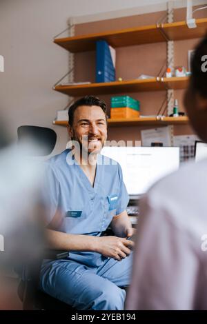 Glücklicher männlicher Arzt in blauen Peelings, der mit dem Patienten spricht, während er im medizinischen Klinikzimmer sitzt Stockfoto
