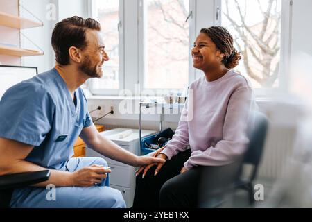 Glücklicher männlicher Arzt tröstet junge Frauen mit der Hand auf dem Knie, während sie im Untersuchungsraum sitzen Stockfoto
