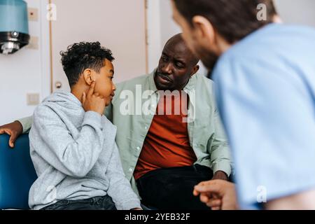 Junge zeigt Ohrenprobleme zum Arzt, während er neben dem Vater im medizinischen Untersuchungsraum sitzt Stockfoto