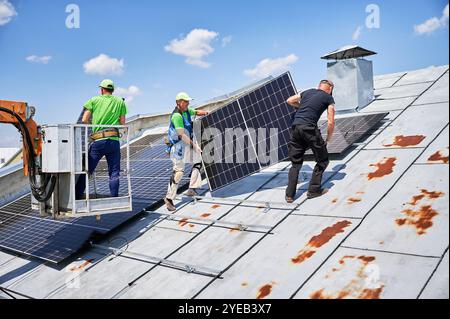 Arbeiter, die Solarpaneele auf dem Metalldach des Hauses mit Hilfe eines Kranaufzugs bauen. Zwei Monteure tragen Photovoltaik-Solarmodul im Freien. Konzept der Erzeugung erneuerbarer Energien. Stockfoto