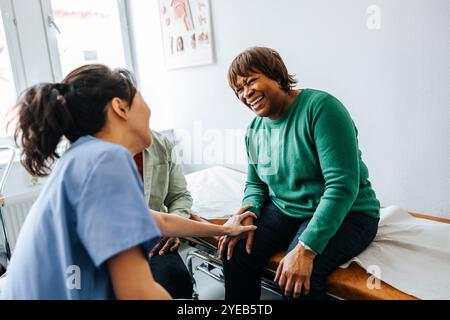 Glückliche ältere Frau, die während des Besuchs mit weiblicher Ärztin im Krankenhaus auf dem Untersuchungstisch sitzt Stockfoto