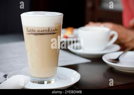 Hohes Glas Latte Macchiato mit Crema und Untertasse, weiße Tasse Kaffee, Kuchen und weibliche Hände im Hintergrund, dunkler Holztisch. Nahaufnahme von Coff Stockfoto