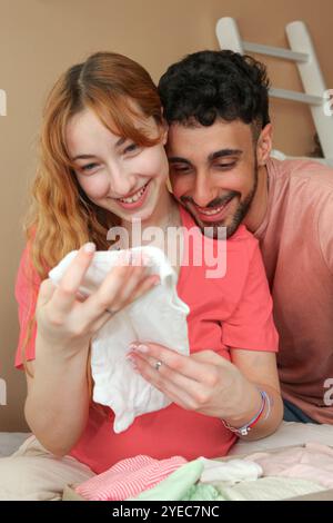 Junge fröhliche schwangere Frau und ihr Mann schauen sich zu Hause ein Paket mit Babykleidung an Stockfoto