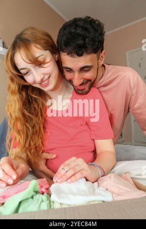 Junge fröhliche schwangere Frau und ihr Mann schauen sich zu Hause ein Paket mit Babykleidung an Stockfoto