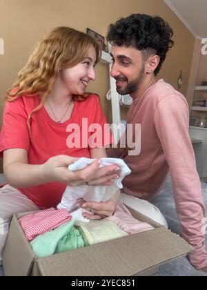 Junge fröhliche schwangere Frau und ihr Mann schauen sich zu Hause ein Paket mit Babykleidung an Stockfoto