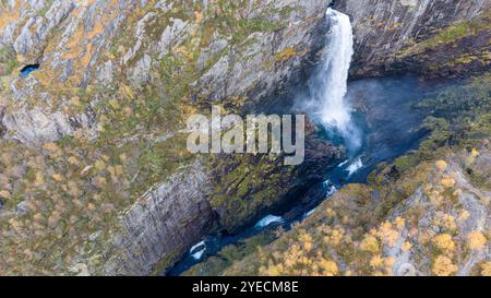 Blick aus der Vogelperspektive auf einen atemberaubenden Wasserfall, der durch felsige Klippen in einen tiefblauen Pool stürzt, umgeben von üppigem Grün und Herbstlaub. Stockfoto