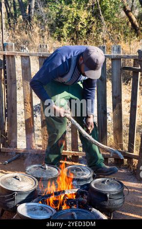 Kessel dreibeiniger Topf im Freien Bulk Cooking, afrikanischer Mann Straßenverkäufer mit kleinem Geschäft, der das Feuer in der Außenküche macht Stockfoto