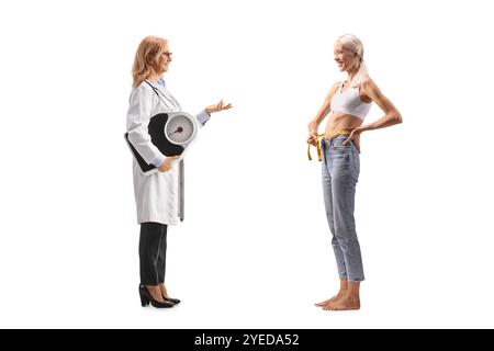 Profilaufnahme einer Ärztin in voller Länge, die eine Waage hält und mit einer schlanken Patientin spricht, isoliert auf weißem Hintergrund Stockfoto
