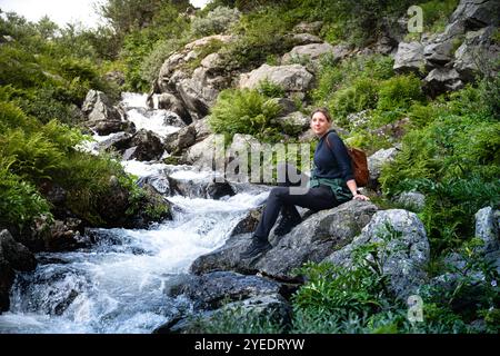 Eine Wandererin macht eine Pause, sitzt auf einem Felsen neben dem rauschenden Fluss Silverfallet in Dalarna Schweden und genießt einen Moment in der lebhaften grünen Natur Stockfoto