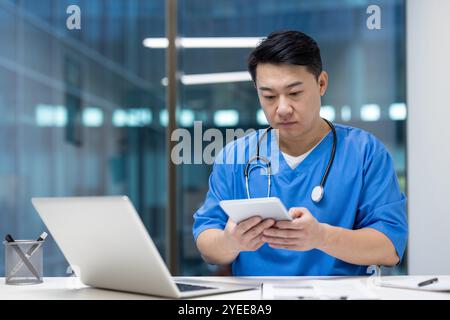 Asiatischer Gesundheitsexperte in Peelings mit Tablet im modernen Büro. Eine ernsthafte Fokussierung auf Technologie erhöht die Effizienz in der medizinischen Umgebung. Laptop, Stethoskop sichtbar, Förderung des medizinischen Personals. Stockfoto