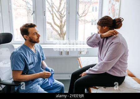 Junge Frau, die Probleme erklärt, während männlicher Arzt Handschuhe im Untersuchungsraum trägt Stockfoto