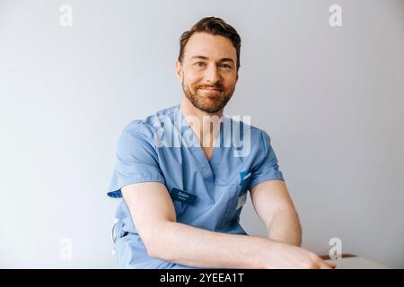 Porträt eines lächelnden männlichen Arztes, der vor der weißen Wand blaue Peelings trägt Stockfoto