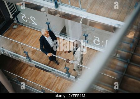 Hochwinkelansicht eines Geschäftsmannes mit Glatze, der mit einem älteren männlichen Kollegen spricht, während er am Glasgeländer steht Stockfoto