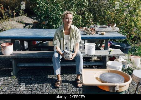 Porträt einer Handwerkerin, die Tassen hält, während sie auf einer Bank neben dem Tisch sitzt Stockfoto
