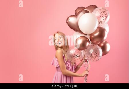 Partyzeit. Charmante blonde Frau in stilvollem Kleid hält Geburtstag Ballons über rosa Studio Hintergrund, kopieren Raum Stockfoto