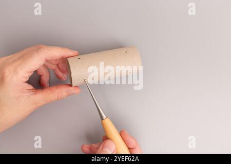 Die Hand hält das Rohr für die Rolle aus Toilettenpapier, während die andere Hand ein Ampelwerkzeug mit einem Holzgriff und einer Metallspitze verwendet, um ein Loch zu erzeugen, ste Stockfoto