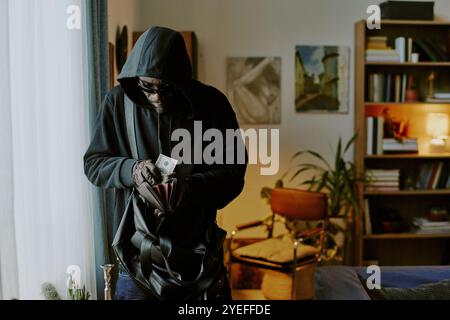 Afroamerikanischer Dieb in Handschuhen, der Ledergeldbörse voll Geld hält und sie herausnimmt, während er die Wohnung raubt Stockfoto