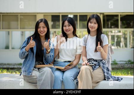 Eine Gruppe fröhlicher und freundlicher asiatischer Studentinnen sitzt zusammen auf einer Bank in ihrem College Park, lächelt und zeigt dem Daumen nach oben Stockfoto