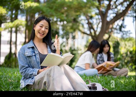 Eine schöne asiatische Studentin in lässiger Kleidung sitzt auf dem Gras in ihrem Campus-Park, liest, zeigt mit dem Finger und lächelt in die Kamera. Stockfoto