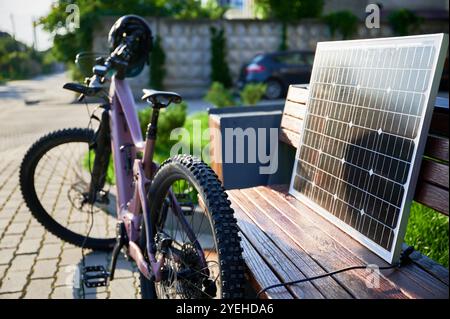 Rosafarbenes E-Mountainbike neben einer Holzbank, verbunden mit Solarmodul zum Laden. Das Fahrrad, mit Helm am Lenker, ruht auf einem gepflasterten Bereich, umgeben von Grün und Gras. Stockfoto