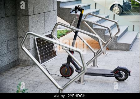 Elektroroller parkt in moderner städtischer Umgebung neben dem Solarmodul auf dem Boden. Elegante graue Steinmauern und Treppen mit Metallgeländern. Nachhaltige Energielösung für den städtischen Pendelverkehr. Stockfoto