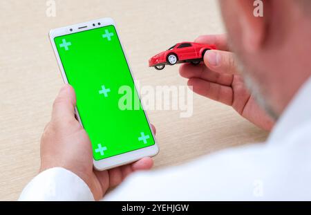 Mann mit grünem Bildschirm, leerem Telefon in vertikaler Position und rotem Spielzeugauto. Anwendung für Verkauf und Miete von Autos Stockfoto