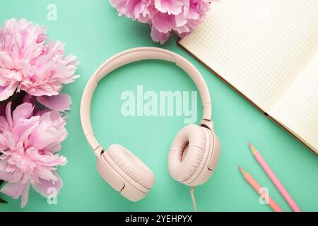 Moderne pinkfarbene Kopfhörer mit Blumen-Pfingstrosen und Notizbuch auf mintfarbenem Hintergrund. Draufsicht Stockfoto