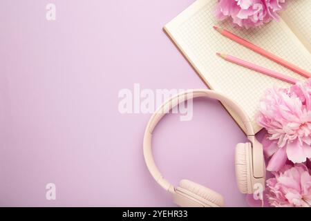 Moderne pinkfarbene Kopfhörer mit Blumen-Pfingstrosen und Notizbuch auf violettem Hintergrund. Draufsicht Stockfoto