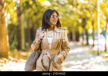 Eine Frau, die einen braunen Trenchcoat und eine Sonnenbrille trägt, geht den Bürgersteig hinunter. Sie lächelt und genießt ihren Tag. Herbst- oder Winterkonzept im ci Stockfoto