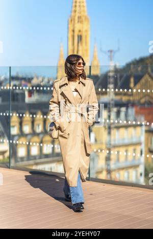 Eine Frau mit einem braunen Trenchcoat und einer blauen Jeans geht auf eine Brücke. Die Skyline der Stadt im Hintergrund ist sichtbar, und die Frau trägt eine Handtasche Stockfoto