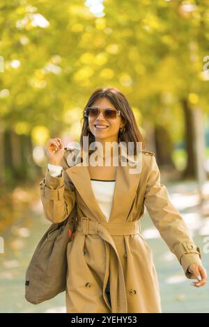 Eine Frau, die einen braunen Trenchcoat und eine Sonnenbrille trägt, geht den Bürgersteig hinunter. Sie lächelt und hält eine Handtasche. Herbst- oder Winterkonzept in der Stadt Stockfoto