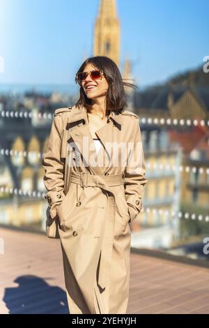 Eine Frau mit einem braunen Trenchcoat lächelt und geht auf einer Brücke. Das Bild hat eine warme und fröhliche Stimmung, mit dem Lächeln der Frau und dem hellen bl Stockfoto