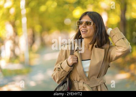 Eine Frau, die einen braunen Mantel und eine Sonnenbrille trägt, läuft einen Weg entlang. Sie lächelt und genießt ihren Spaziergang. Herbst- oder Winterkonzept in der Stadt Stockfoto