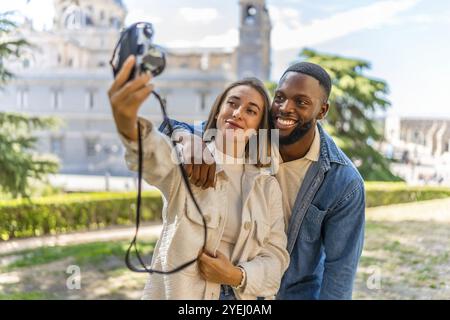 Glückliches Multi-ethnisches Paar, das Selfie mit Digitalkamera in einem Park mit Blick auf die Innenstadt macht Stockfoto