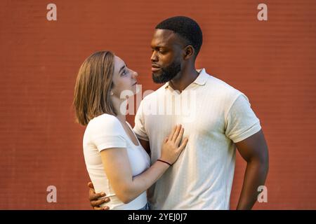 Gemischtes Paar, das sich umarmt und an der roten Ziegelmauer ansieht Stockfoto