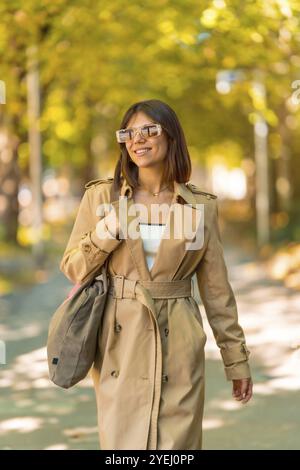 Eine Frau, die einen braunen Trenchcoat und eine Sonnenbrille trägt, geht den Bürgersteig hinunter. Sie trägt eine braune Handtasche. Herbst- oder Winterkonzept in der Stadt Stockfoto