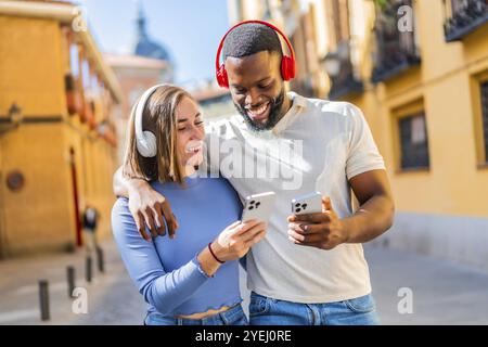 Glückliches, multiethnisches Paar, das in der Stadt unterwegs ist und Musik hört Stockfoto