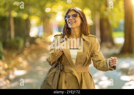 Eine Frau, die einen braunen Trenchcoat und eine Sonnenbrille trägt, läuft einen Weg entlang. Sie lächelt und genießt ihren Spaziergang. Herbst- oder Winterkonzept im Stockfoto